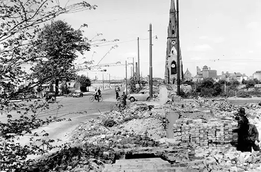 Blick auf eine vom Krieg vollständig zerstörte Strasse und Häuser. Ein Kirchturm im Hintergrund.