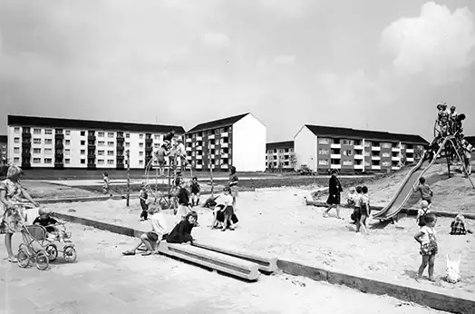 Kinder auf einem Sandspielplatz. Im Hintergrund Wohnblöcke. Schwarz-weiss