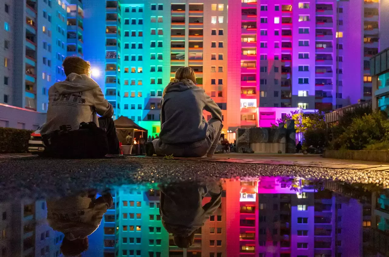 Hochhäuser mit bunten Lichtern angeleuchtet bei Nacht vor denen zwei Personen sitzen.