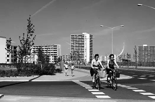 Zwei radfahrende Personen auf einer Strasse. Im Hintergrund Hochhaus-Wohnblöcke. Schwarz-weiss