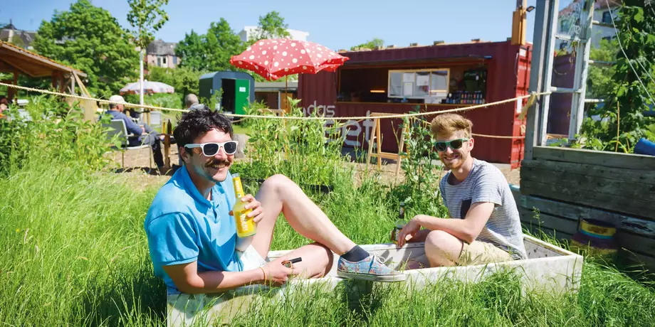 Zwei Männer sitzend in einem Kasten, der im Gras steht vor einem Containerkiosk