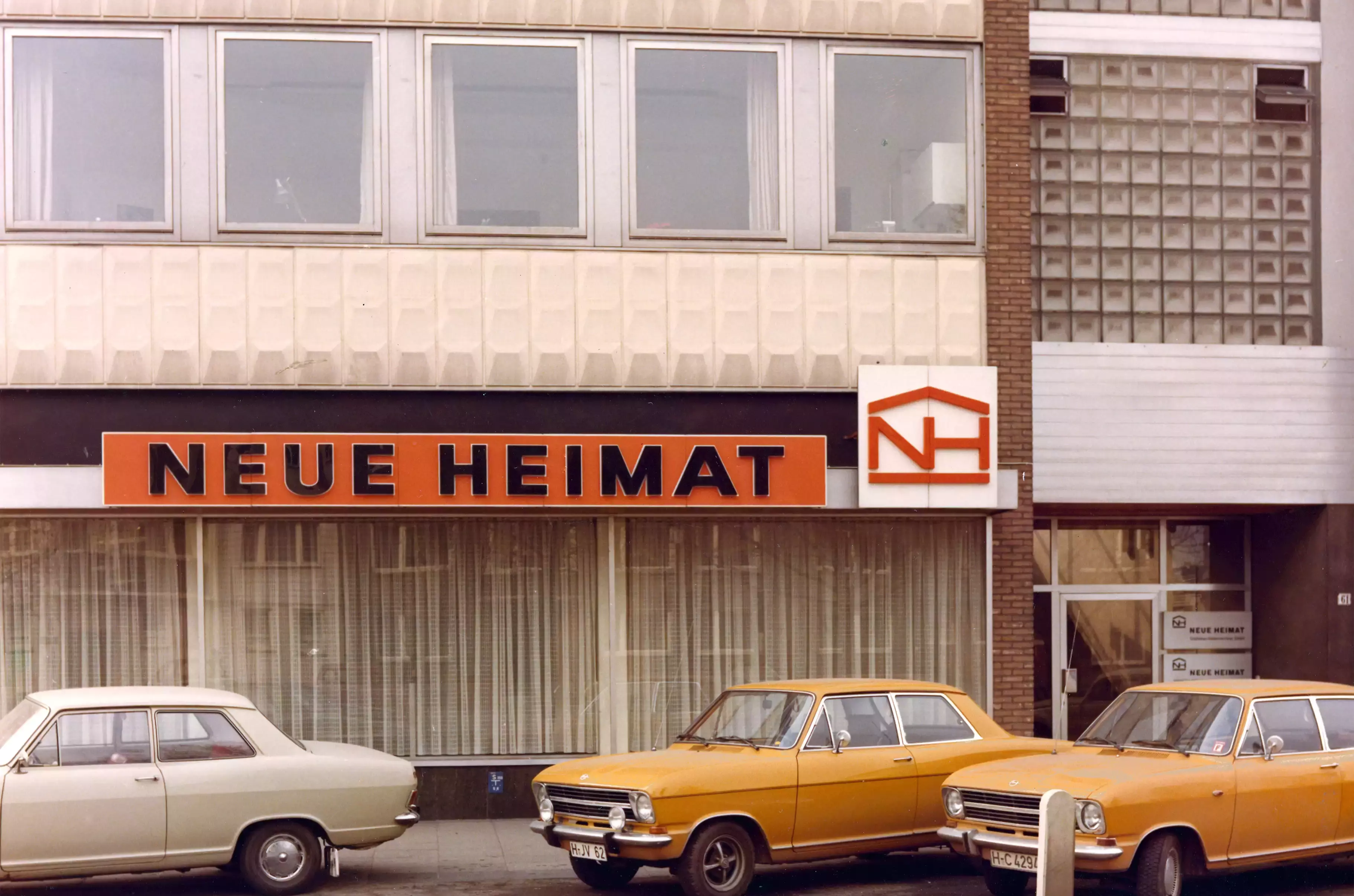Gelbe Autos vor einem Büro mit orangem Schild "Neue Heimat"